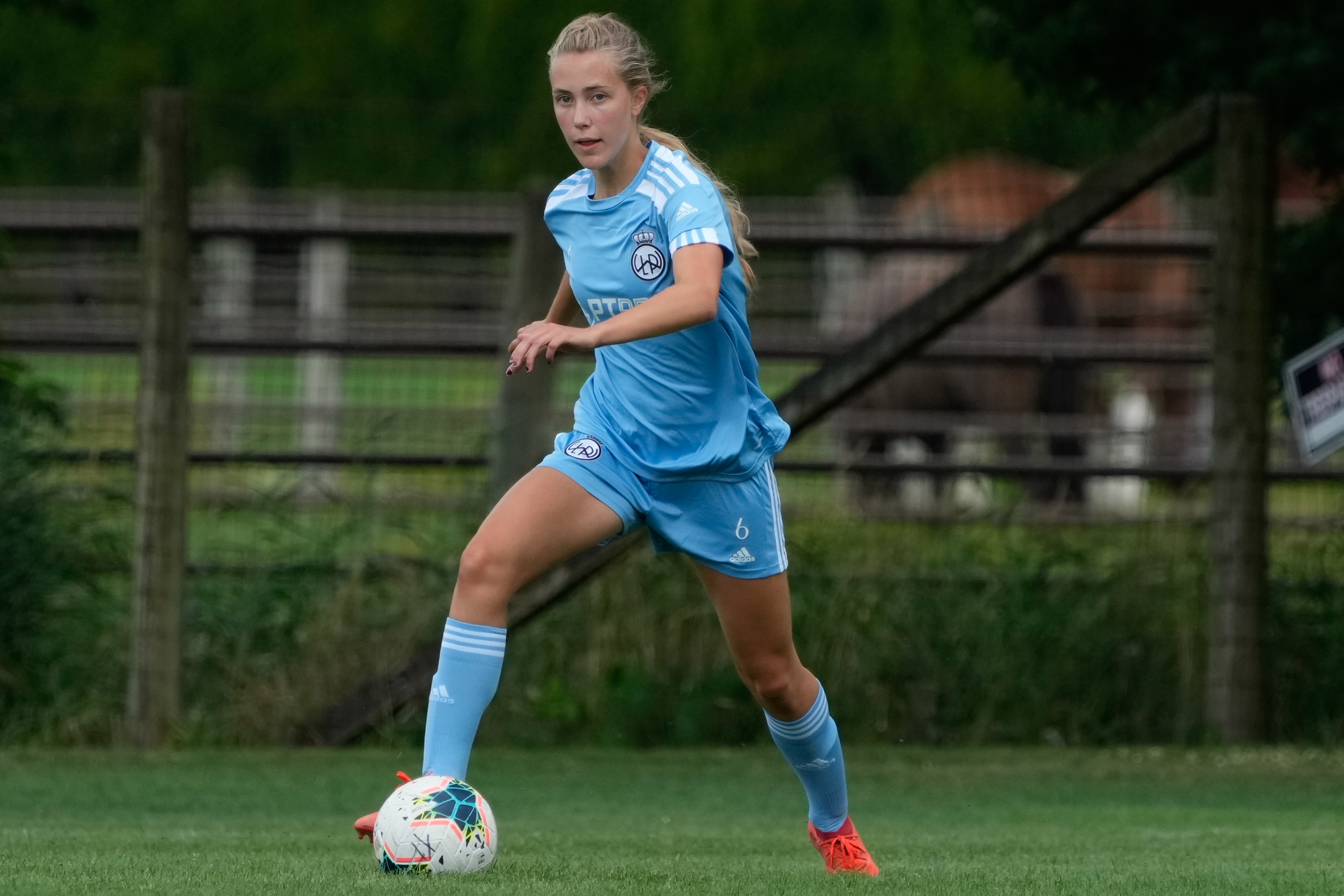 Female player wearing a light blue Adidas uniform running with a soccer ball during a game.